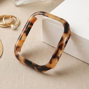 May include: A tortoiseshell-patterned Apple Watch case sits near a white box with "Apple Watch Series 9" printed on it. A gold necklace, rings, and a perfume bottle are also in the image.