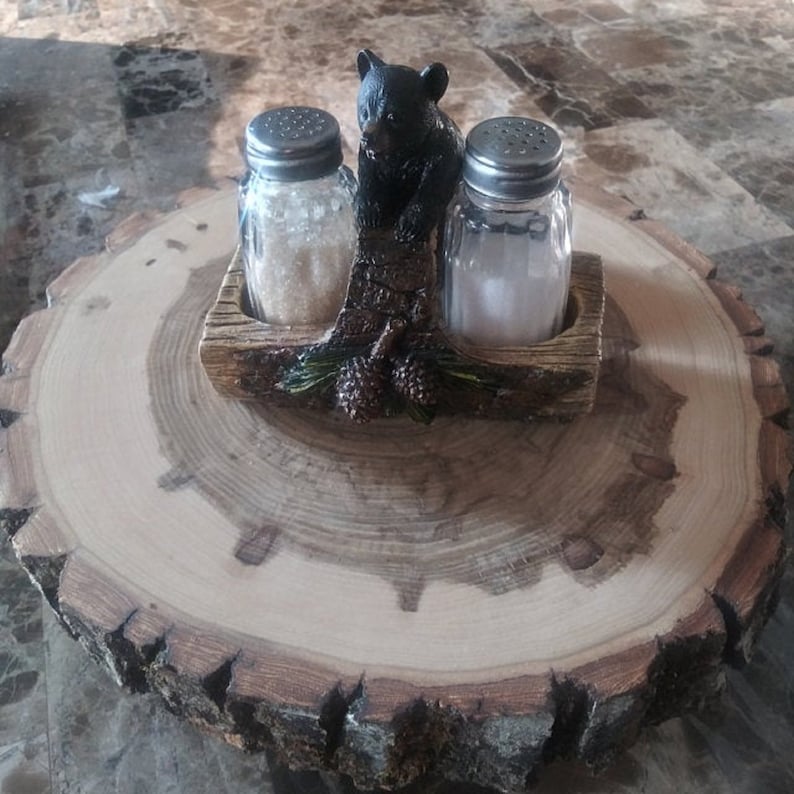 May include: Salt and pepper shakers in a wooden holder with a black bear figurine on top. The holder is sitting on a wooden slice.