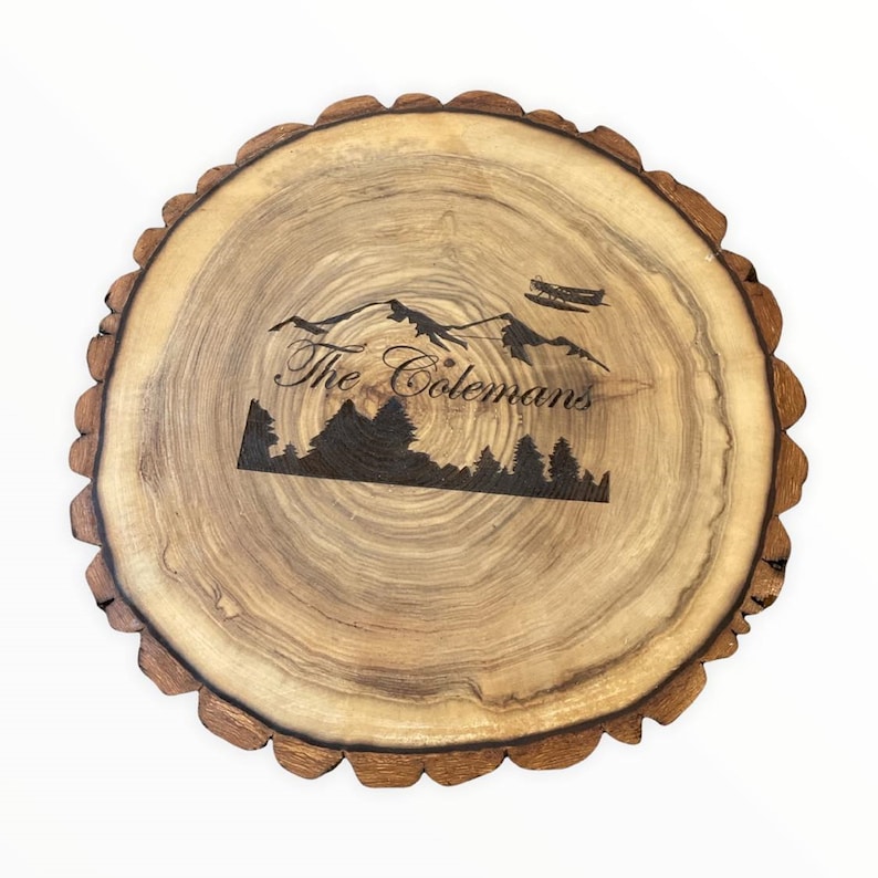 May include: A round wooden serving tray with a natural wood grain finish. The tray has a rough, uneven edge and features a laser-engraved design of mountains, trees, and a small airplane. The text "The Colemans" is engraved below the design.