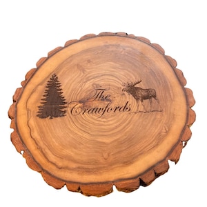 May include: A round wooden slice with a natural bark edge. The wood is a light brown color with darker brown rings. The slice is engraved with a moose, a pine tree, and the text "The Crawfords".
