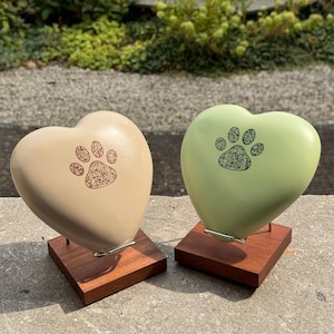 Può includere: Due pietre commemorative a forma di cuore, una beige e una verde chiaro, ciascuna con un design a zampa scura con un codice QR integrato. Entrambe le pietre sono montate su basi quadrate in legno con supporti in filo metallico sottile.