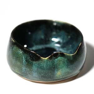Op de afbeelding: Een kleine, ronde keramische kom met een diepe, glanzende glazuurlaag in tinten donker bosgroen, teal en zwart. De rand van de kom heeft aan één kant een duidelijke, subtiele inkeping en de binnenzijde heeft een gladde, reflecterende afwerking.