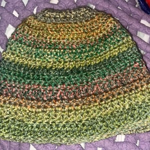 Puede incluir: Un gorro tejido a ganchillo a mano con un patrón de hilo jaspeado con rayas horizontales en tonos verde bosque, oliva, lima y sutiles toques de naranja óxido. El gorro tiene una corona redondeada y un patrón de punto texturizado en toda la tela.