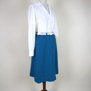 May include: A white long-sleeve blouse with a collar and button-down front is paired with a teal skirt. A white belt cinches the waist. The outfit is displayed on a wooden dress form.