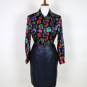 Könnte beinhalten: Ein schwarzes Langarmhemd mit einem farbenfrohen geometrischen Muster aus roten, grünen, blauen und goldenen Quadraten. Kombiniert mit einem schwarzen Bleistiftrock aus Leder. Das Outfit wird auf einer Holzpuppe präsentiert.