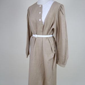 May include: A tan-colored dress with long sleeves and a white collar is displayed on a mannequin. The dress features a white belt and three white buttons down the front. The dress is made of a linen-like material.