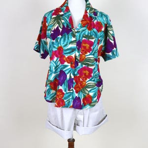 May include: A short-sleeved shirt with a colourful floral pattern in red, purple, and green on a turquoise base. The shirt is worn with white shorts. The clothing is displayed on a mannequin.