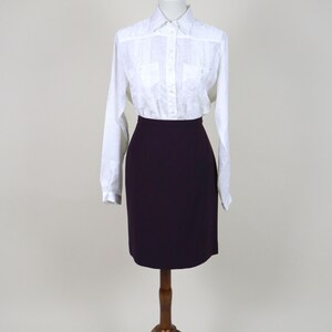 Op de afbeelding: Een witte blouse met subtiel patroon en een donkerpaarse potloodrok worden gepresenteerd op een houten mannequin. De blouse heeft lange mouwen en twee borstzakken. De rok is knielang.