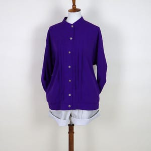 Peut inclure: Une veste violette vibrante avec un col mandarin et des manches longues. La veste présente des plis verticaux sur le devant et est fermée par des boutons argentés. Elle est présentée sur un mannequin avec un short gris clair.