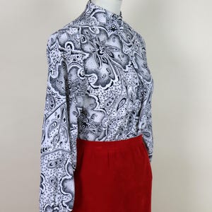 May include: A long-sleeved blouse with a high collar and a red skirt. The blouse features a black and white paisley pattern. The skirt is a solid, vibrant red color. The clothing is displayed on a mannequin.
