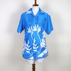 May include: A blue short-sleeved shirt featuring a white floral design, including bird-of-paradise flowers and tropical leaves. The shirt is displayed on a mannequin. It has a classic collar and button-down front.