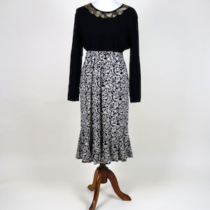 May include: A black long-sleeved top with gold trim around the neckline and a black and white patterned skirt. The skirt has a flared hem. The outfit is displayed on a wooden dress form.