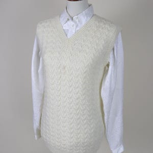 May include: Cream-colored, V-neck sweater vest with a textured, patterned design. The vest is layered over a white collared shirt with long sleeves. The outfit is displayed on a mannequin.