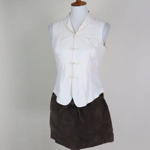 May include: A white sleeveless top with a mandarin collar and decorative frog closures, paired with a brown suede mini skirt. The outfit is displayed on a mannequin against a neutral background, showcasing a vintage or retro style.