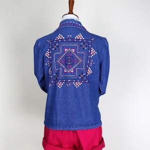 Peut inclure: Veste en jean bleue avec un motif géométrique brodé dans le dos. La broderie présente un motif carré avec des accents blancs, roses, violets et jaunes. La veste est associée à un short rouge.