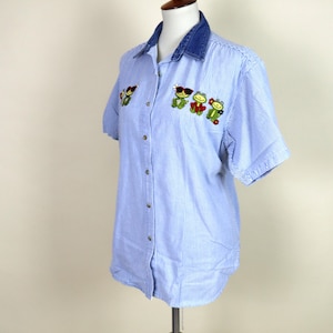 May include: Blue and white striped short-sleeve shirt with a denim collar. The shirt features embroidered frog designs wearing sunglasses and flowers. The shirt is button-down and displayed on a mannequin.