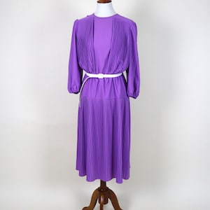 Könnte beinhalten: Ein vintage lila Kleid mit langen Ärmeln und einem plissierten Oberteil und Rock. Das Kleid ist mit einem weißen Gürtel in der Taille zusammengezogen. Das Kleid wird auf einer hölzernen Schneiderpuppe präsentiert.