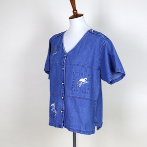 May include: Blue denim short-sleeve shirt with a V-neck and button-up front. The shirt features embroidered flamingo and tropical plant designs on the pockets and upper chest. The shirt is displayed on a mannequin.