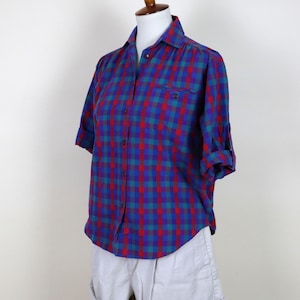 May include: A button-down plaid shirt in red, blue, and teal, with rolled-up sleeves, displayed on a mannequin. The shirt has a chest pocket and is paired with light-coloured shorts.