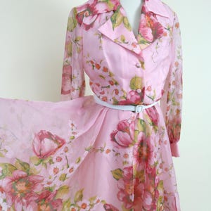 Puede incluir: Vestido rosa vintage con estampado floral. El vestido presenta un cuello, mangas largas y un cinturón blanco. El diseño floral incluye flores rosas, rojas y verdes. El vestido se exhibe en un maniquí.