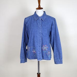 Peut inclure: Chemise en jean bleu avec des manches longues et une fermeture boutonnée. La chemise présente des motifs floraux brodés en rose, violet et bleu près de l'ourlet. La chemise est présentée sur un mannequin.
