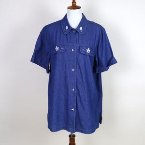 May include: A short-sleeved, dark blue denim shirt on a mannequin. The shirt has a collared neckline with white embroidered accents and leaf designs on the chest pockets. It is buttoned down the front.