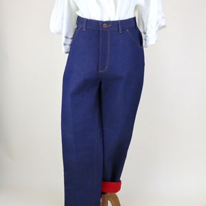 Puede incluir: Pantalones vaqueros azul oscuro de cintura alta con costuras naranjas. Los pantalones están doblados en el bajo, revelando un forro rojo. Una blusa blanca con detalles de encaje es visible en la parte superior.