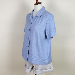 May include: A light blue short-sleeved button-down shirt with a subtle striped pattern. The shirt is displayed on a mannequin with beige shorts. The shirt has a classic collar and a pleated hemline.