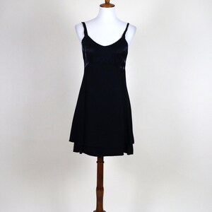 May include: A black slip dress with spaghetti straps and a V-neckline. The dress is displayed on a white mannequin atop a wooden stand. The dress has a slightly flared skirt and a satin-like bodice.