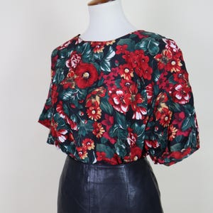 May include: A floral print blouse with a black background, featuring red, white, and yellow flowers and green leaves. The blouse is paired with a black leather skirt, displayed on a mannequin.