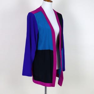 May include: A color-blocked jacket in shades of purple, blue, and black, displayed on a mannequin. The jacket features long sleeves and a contrasting magenta trim along the edges. The design is a bold, geometric pattern.