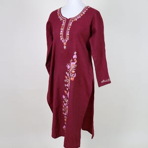 Puede incluir: Vestido túnica burdeos con mangas largas, exhibido en un maniquí. El vestido presenta intrincados bordados florales en tonos rosa, naranja y blanco a lo largo del escote y en la parte delantera. Tiene una abertura lateral y está hecho de tela suave.