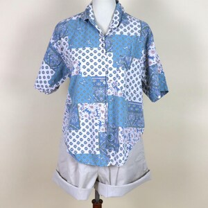 May include: A short-sleeved shirt with a patchwork design in blue and white, featuring floral and geometric patterns. The shirt is paired with light beige shorts, displayed on a mannequin. The shirt has a collar.