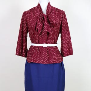 May include: A burgundy blouse with a white dotted pattern and a bow tie collar, paired with a white belt and a blue pencil skirt. The blouse has three-quarter sleeves. The outfit is displayed on a mannequin.