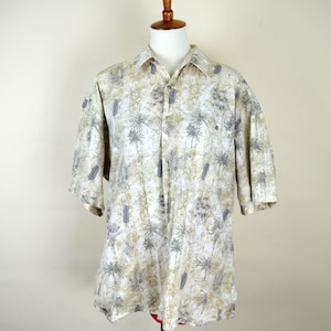 Może przedstawiać: Koszula z krótkim rękawem zapinana na guziki z tropikalnym nadrukiem. Koszula ma białe tło z wzorem palm i szarymi i beżowymi akcentami. Koszula jest prezentowana na drewnianym manekinie.
