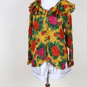 Puede incluir: Una blusa con estampado floral y cuello con volantes, exhibida en un maniquí. La blusa presenta un fondo amarillo con rosas rojas y rosas y hojas verdes. Tiene mangas largas y se combina con pantalones cortos blancos.