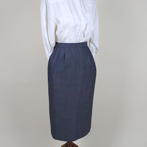 Puede incluir: Una camisa blanca abotonada con un sutil estampado floral y una falda lápiz azul oscuro. La falda llega a la rodilla y tiene un corte entallado. La ropa se exhibe en un maniquí de madera.