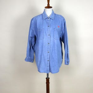 May include: Light blue denim shirt with long sleeves, two chest pockets, and a button-down front. The shirt features a small embroidered floral design on the left chest. The shirt is displayed on a wooden mannequin.