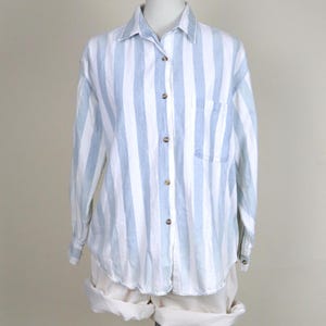 May include: A light blue and white striped button-down shirt with a single chest pocket. The long-sleeved shirt is displayed on a mannequin with rolled-up white shorts. The shirt has a classic collar and wooden buttons.
