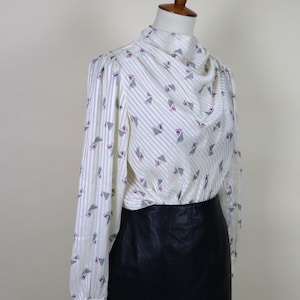 Puede incluir: Una blusa vintage marfil con rayas verticales y patrones geométricos triangulares en morado, verde azulado y negro. La blusa tiene un cuello drapeado y mangas largas. Combinada con una falda de cuero negra.