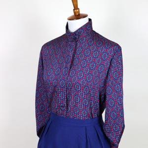 May include: A vintage blouse and skirt set on a mannequin. The blouse has a high collar and a repeating pattern of red, blue, and teal shapes on a dark blue background. The skirt is a solid, deep blue colour.