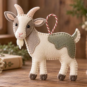 May include: A felt goat ornament featuring a cream-colored body with gray and sage green patches, small black eyes, a pink stitched nose, and a small beard. The goat has two horns and a red and white striped string loop attached to its back.