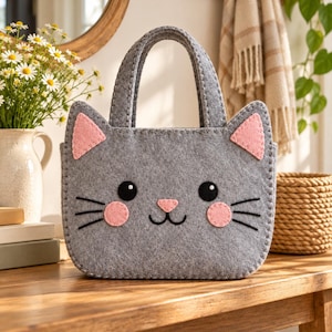 Könnte beinhalten: Eine graue Filztasche in Form eines Katzengesichts mit zwei Tragegriffen, rosa dreieckigen Innenohren, rosa runden Wangen, schwarzen gestickten Augen mit weißen Akzenten, einer rosa Nase und schwarzen gestickten Schnurrhaaren auf der Vorderseite.