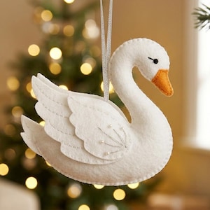 Può includere: Un ornamento a forma di cigno in feltro bianco con un becco arancione brillante, un occhio ricamato nero e ricami in filo argentato sull'ala. Il cigno è appeso a un sottile nastro argentato e presenta ali in feltro sovrapposte con perline.