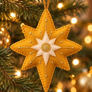 May include: A mustard yellow felt star ornament featuring a smaller white felt star layered on top. The center has a circular gold glitter accent. Gold metallic thread embroidery radiates from the center, and a gold cord loop is attached to the top.