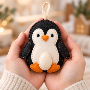 May include: A felt penguin ornament featuring a black body, a white belly and face, and orange feet and beak. The penguin has small black eyes and is constructed with visible stitching along the edges. A beige fabric loop is attached to the top.