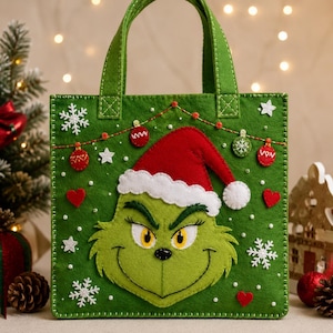 Könnte beinhalten: Eine grüne Filztasche mit einer zentralen Applikation eines grünen Charaktergesichts, das eine rot-weiße Weihnachtsmannmütze trägt. Die Tasche ist mit weißen Filzschneeflocken, roten Herzen und einer Schnur aus kleinen roten und grünen Ornamenten verziert.