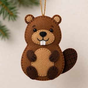 May include: A brown felt beaver ornament featuring a tan belly, dark brown paws, and a textured tail. The beaver has black bead eyes, a black nose, and two white felt teeth. A thin brown string is attached to the top for hanging purposes.