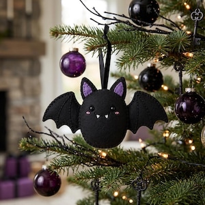 Puede incluir: Un adorno de murciélago de fieltro negro con detalles morados cuelga de un árbol de Navidad. El murciélago tiene una cara sonriente y está rodeado de adornos morados y plateados. El árbol está decorado con adornos en forma de llave y luces.