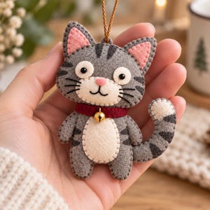 Puede incluir: Un adorno de fieltro con forma de gato gris y blanco, con rayas gris oscuro, orejas rosadas, una pequeña nariz rosa y un collar rojo con un cascabel dorado. El gato tiene ojos y bigotes bordados en negro y un cordón marrón para colgar.